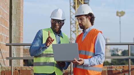 Africa-American-young-male-builder-and-Caucasian-architect-in-hardhats-standing-at-the-roof-of-the-building-site-with-a-laptop-computer-and-talking-about-their-work.